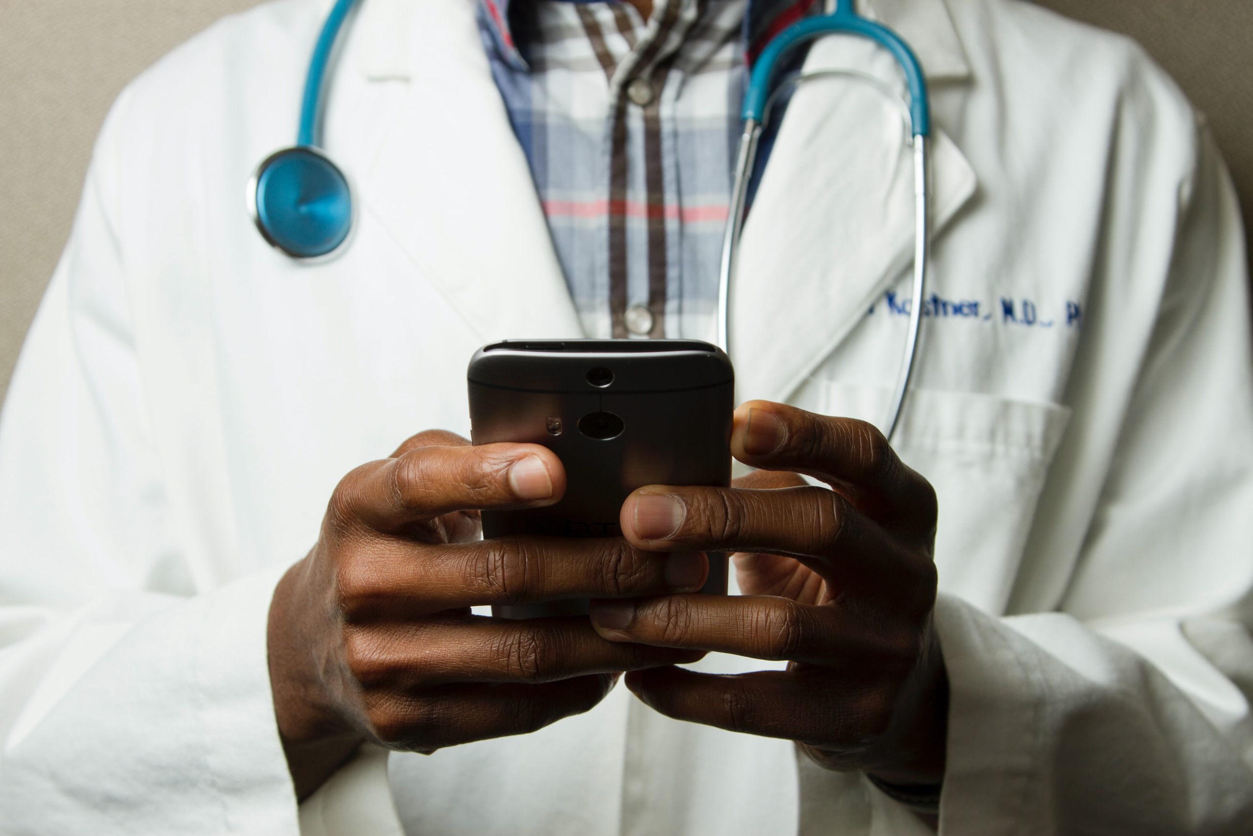 Male doctor wearing a white coat and a stethoscope around his neck holding a smartphone in his hands.