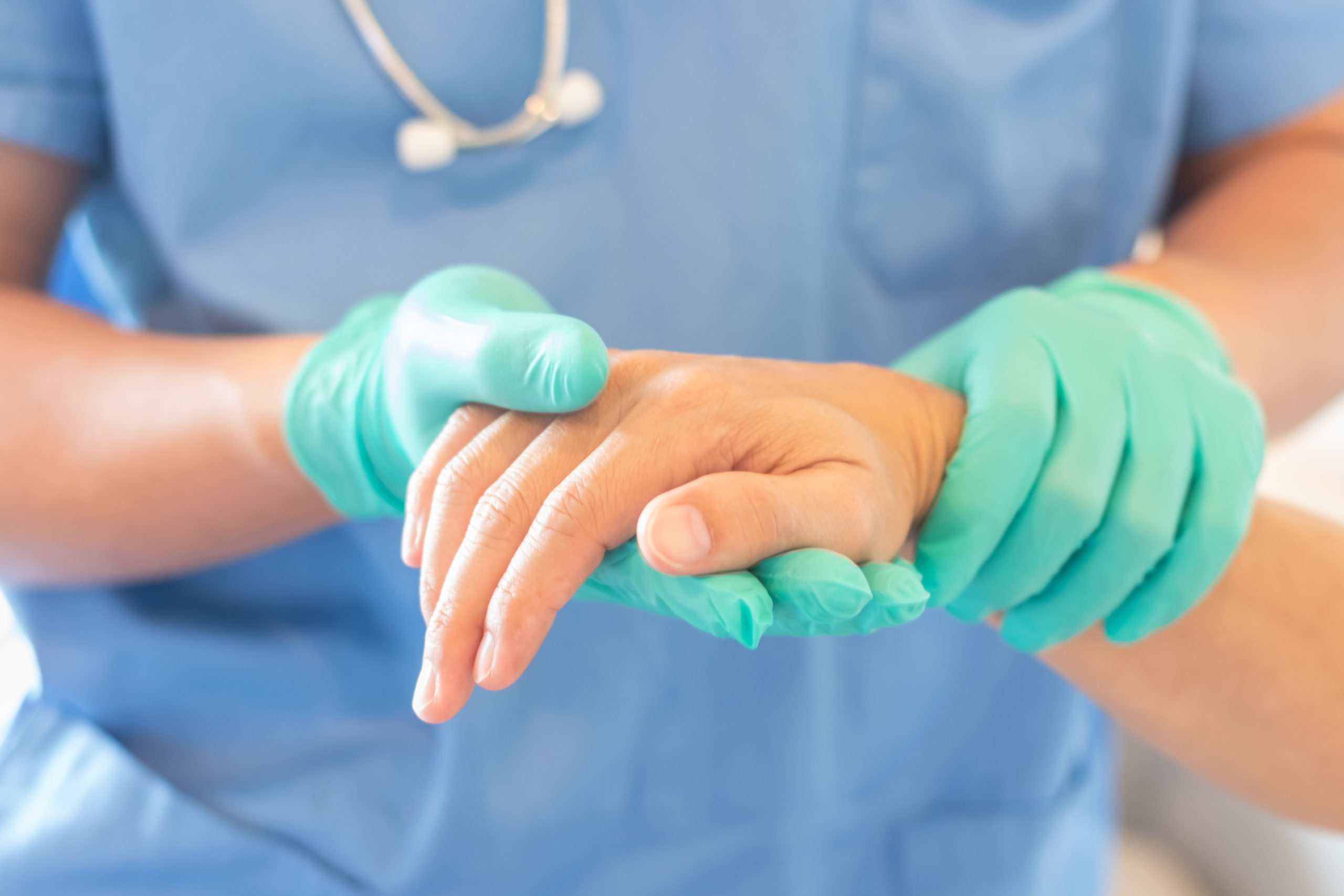 Health professional in a blue uniform wearing green medical gloves holding a patients hand.