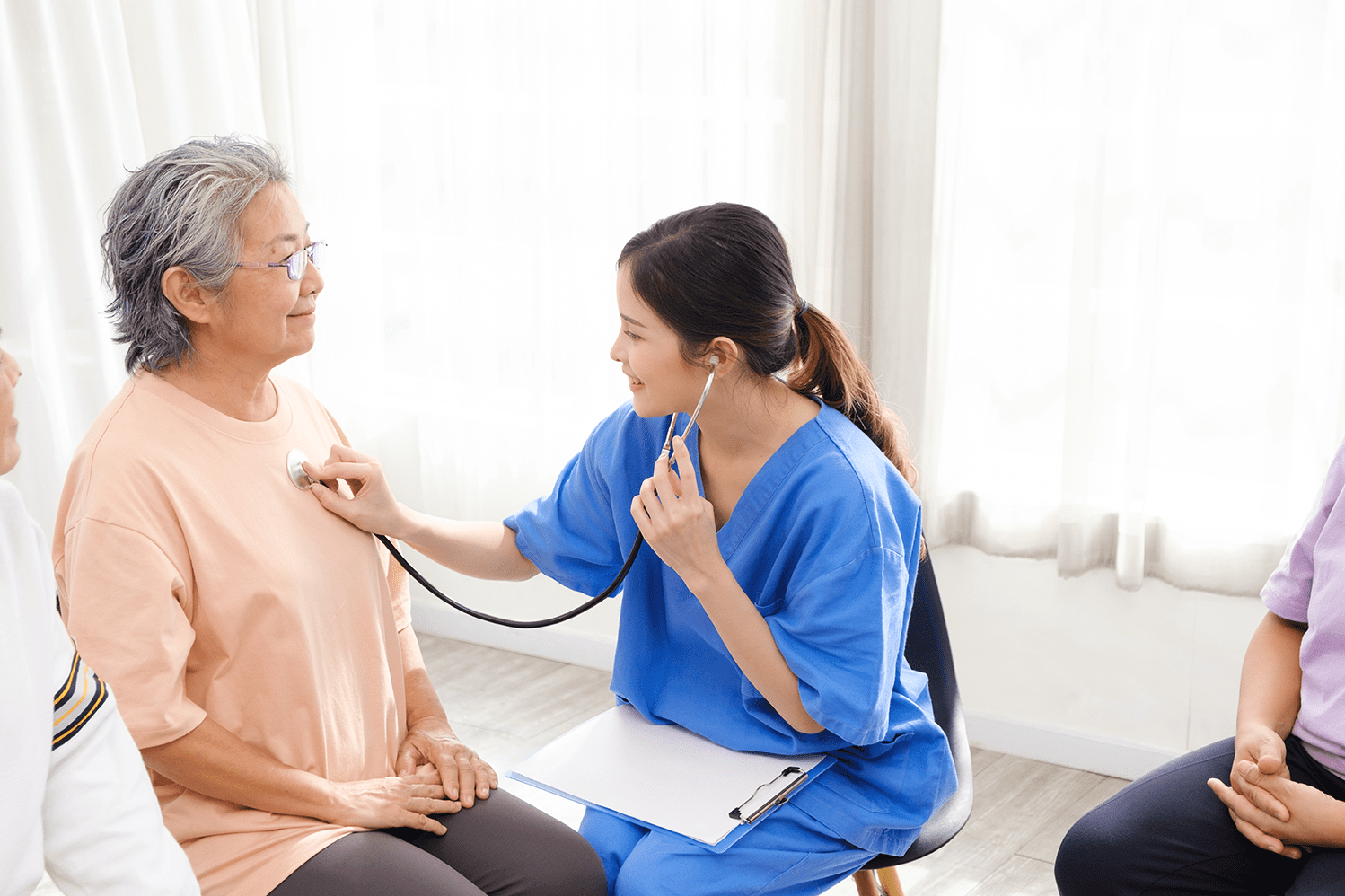 Lady in blue nurse's overalls taking heart beat reading of lady in dusty pink t-shirt using a stethoscope