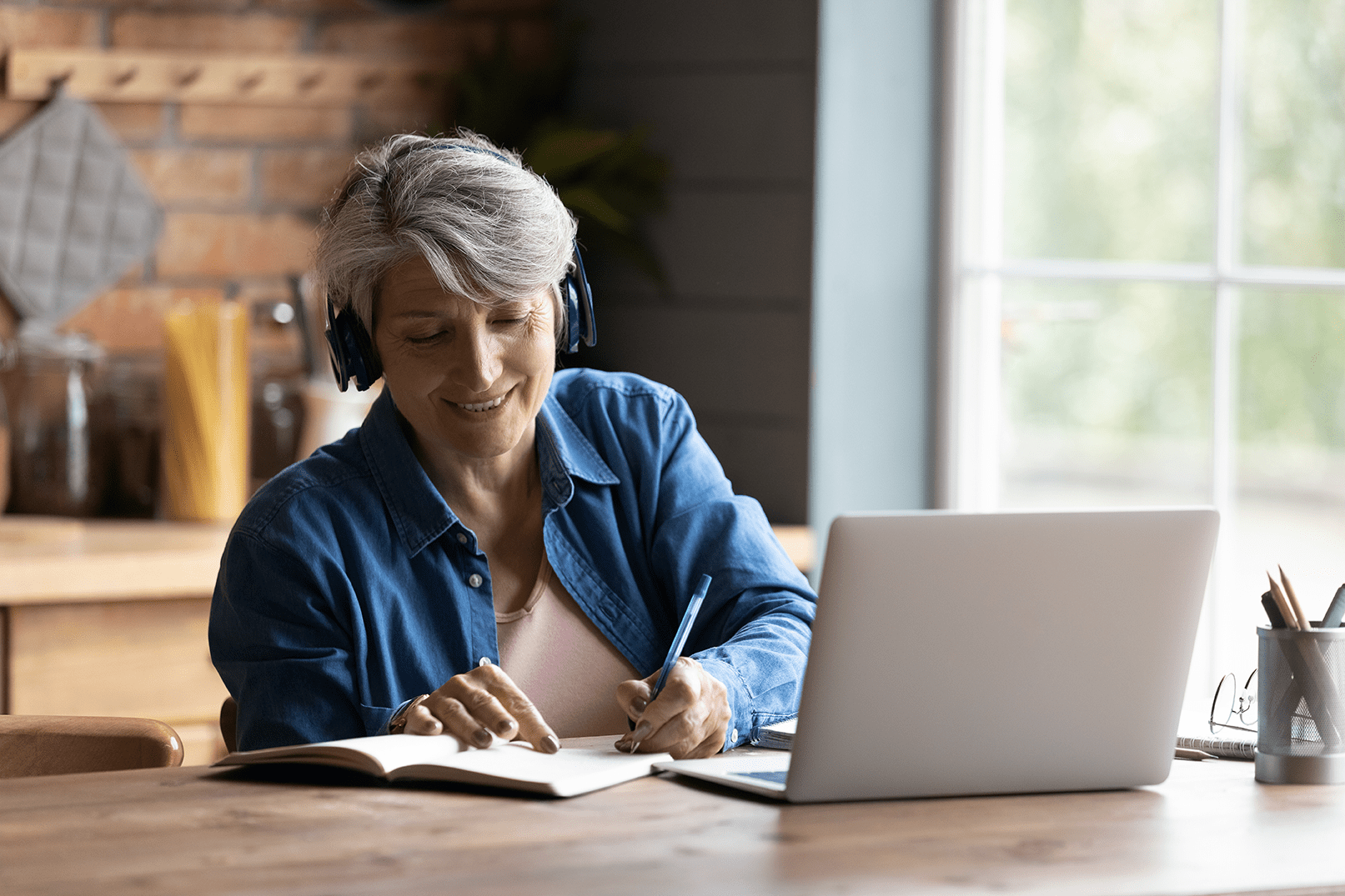 Lady sitting at kitchen table writing on notepad with open laptop in front of her